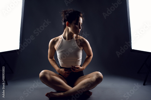 Portrait of young woman athlete in black high-waist panties and white top sitting in lotus position on the floor between two studio flash lights and looking aside. Isolated on grey background