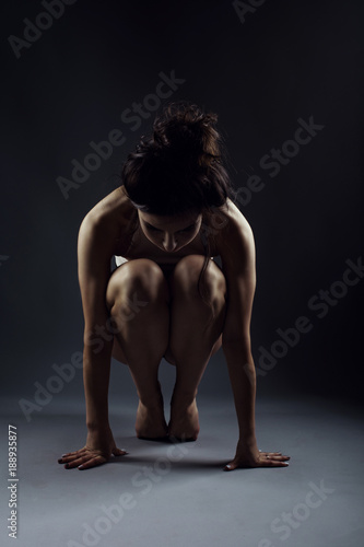 Portrait of young woman athlete sitting on tiptoes in the squatting position on the floor between two studio flash lights with lowered head. Isolated on grey background © obrik