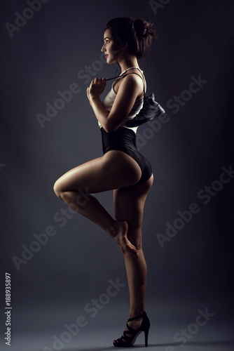 Portrait of young smiling female dancer in black high waist panties and white top standing on one leg in high-heeled shoe, another leg is barefoot and raised. She is holding a boot behind her shoulder © obrik