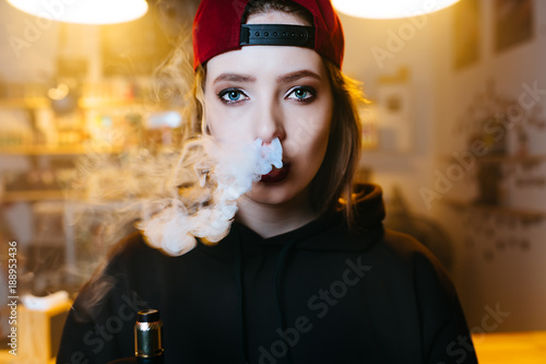 Young pretty woman in red cap smoke an electronic cigarette at the vape shop. Hip-hop style. Closeup. photo