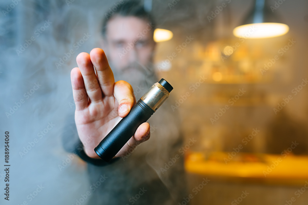 Young beard man show vaping device on his outstretched hand through a cloud of steam. Selective focus. Vaping concept. Copy space. Young beard man show vaping device on his outstretched hand through a cloud of steam. Selective focus. Vaping concept. Copy space.