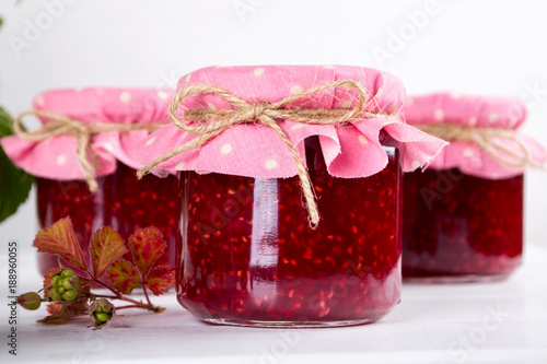 Raspberry jam in glass jar, fresh  ripe raspberry and green leaves on white wooden table