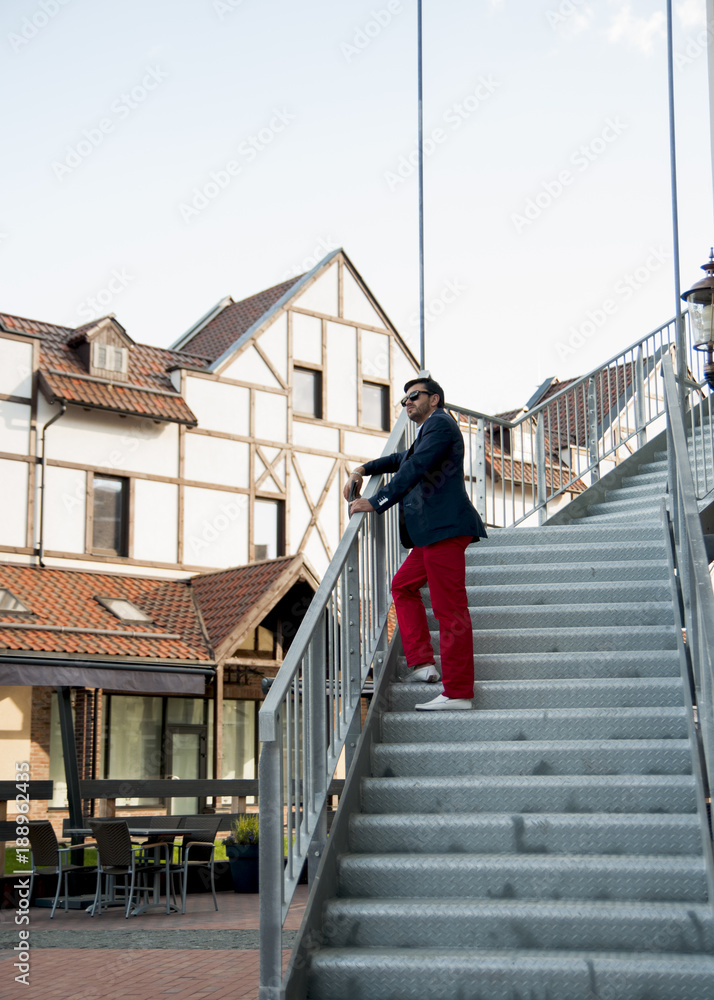 A man in black glasses and red trousers stands on the stairs and looks into the distance