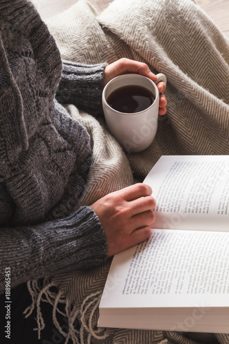 Woman wearing gray woolly cardigan enjoying a mug of tea and reading a book under a cozy blanket