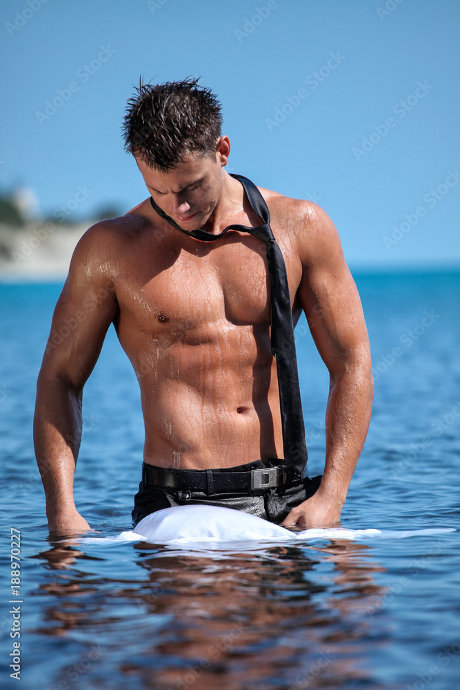 Muscular young sexy wet guy standing in the sea and takes off his business suit with a tie Stock
