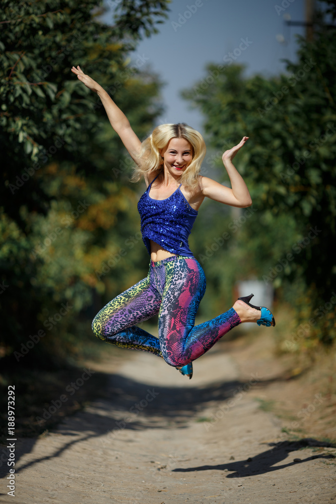 young smiling girl in colored leggings and blue top on heels jumps in the park young smiling girl in colored leggings and blue top on heels jumps in the park