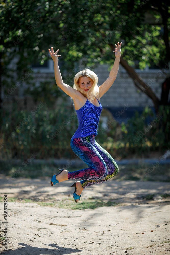 young smiling girl in colored leggings and blue top on heels jumps in the park young smiling girl in colored leggings and blue top on heels jumps in the park