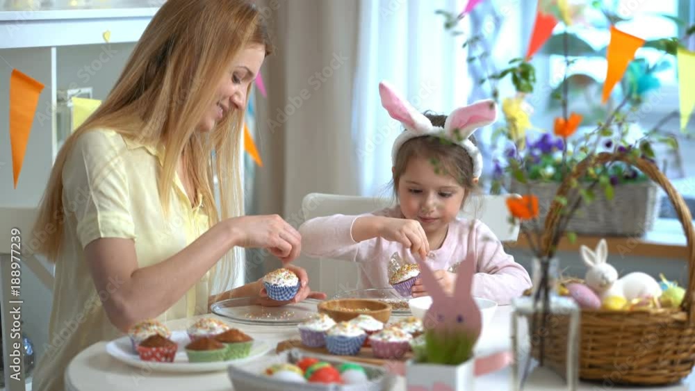 Young mother and her cute little daughter wearing funny rabbit ears are cooking Easter cupcakes sitting at a festive table with basket, eggs and Bunny Young mother and her cute little daughter wearing funny rabbit ears are cooking Easter cupcakes sitting at a festive table with basket, eggs and Bunny