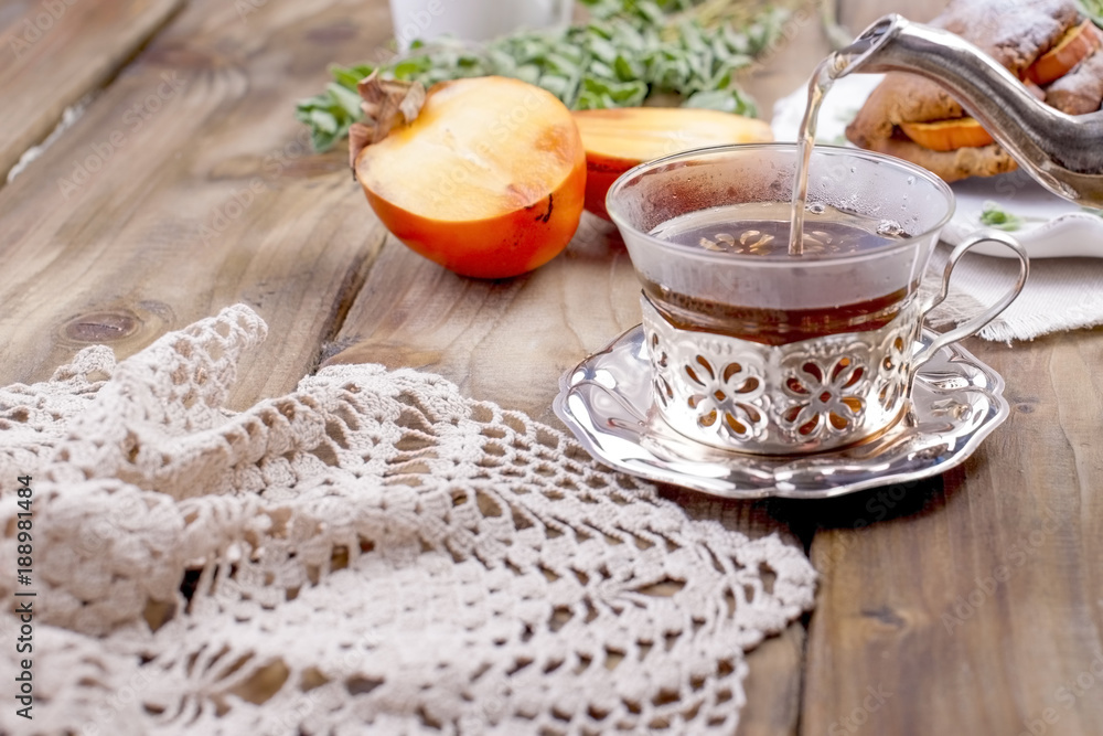 Fototapeta premium Homemade pastries with persimmons, for breakfast. Beautiful glass with tea. Kettle. Wooden table, tablecloth with lace. Free space for text or advertising.