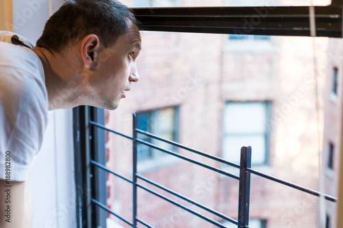 Closeup of young man's face looking outside of small apartment window in New York City NYC urban Bronx, Brooklyn brick housing, guard rail, security bars, checking weather