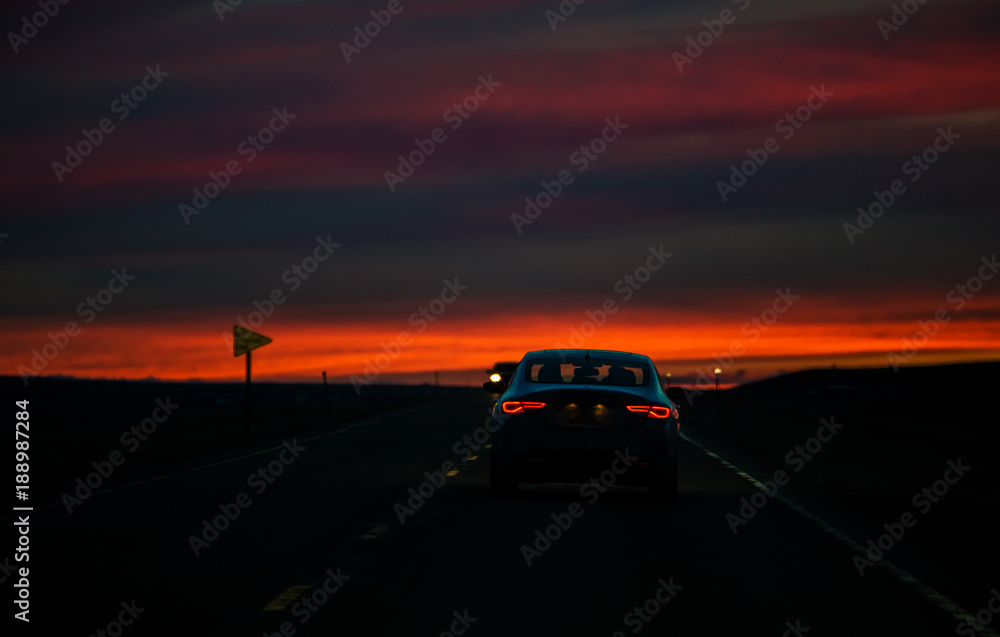 Red taillights of a travelling car on a highway against a colorful sunset in a rural landscape