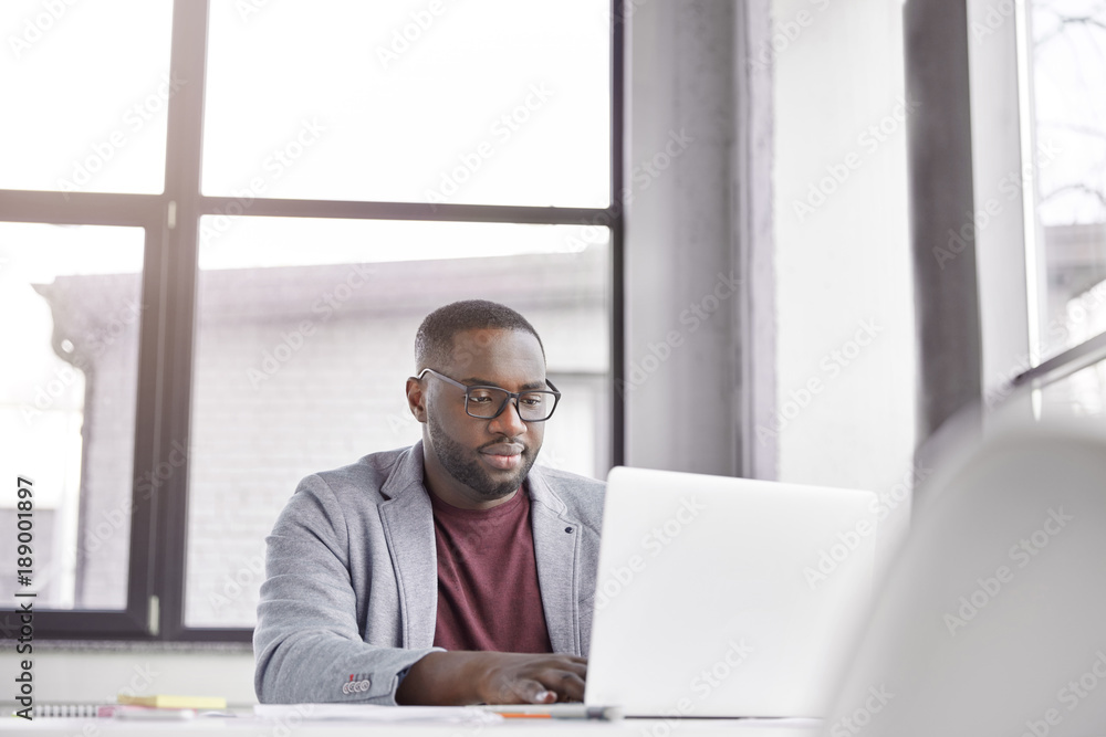 Fototapeta premium Indoor shot of professional black male freelancer works remotely on laptop computer, plans working schedule, keyboards information, focused into screen. Trader analyzes financial market online