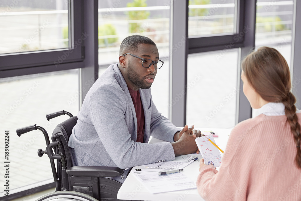 Disabled male boss has dark skin, dressed in formal cothes, analyzes budget receipts and outlays together with female assistant, can`t understand something in graphics, sit over loft office interior Disabled male boss has dark skin, dressed in formal cothes, analyzes budget receipts and outlays together with female assistant, can`t understand something in graphics, sit over loft office interior