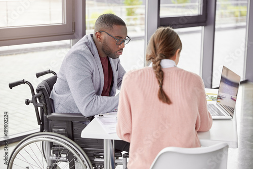 Obraz na plátne Serious dark skinned male invalid in wheelchair collaborates with young female, make business plan together, watch online conference or webinar online, sit in spacious cabinet