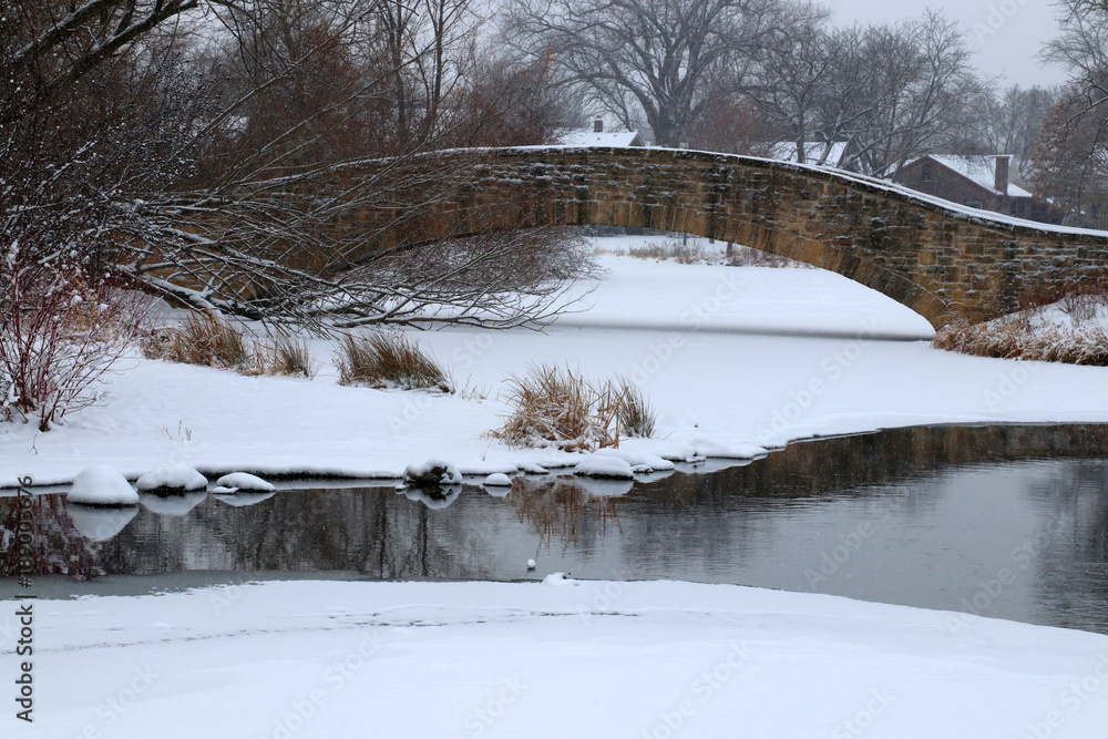 Scenic winter view in a city park. Winter snowy day landscape with an old style brick bridge during snowfall. Tenney Park in the city of Madison, the capital of Wisconsin, Midwest USA.
