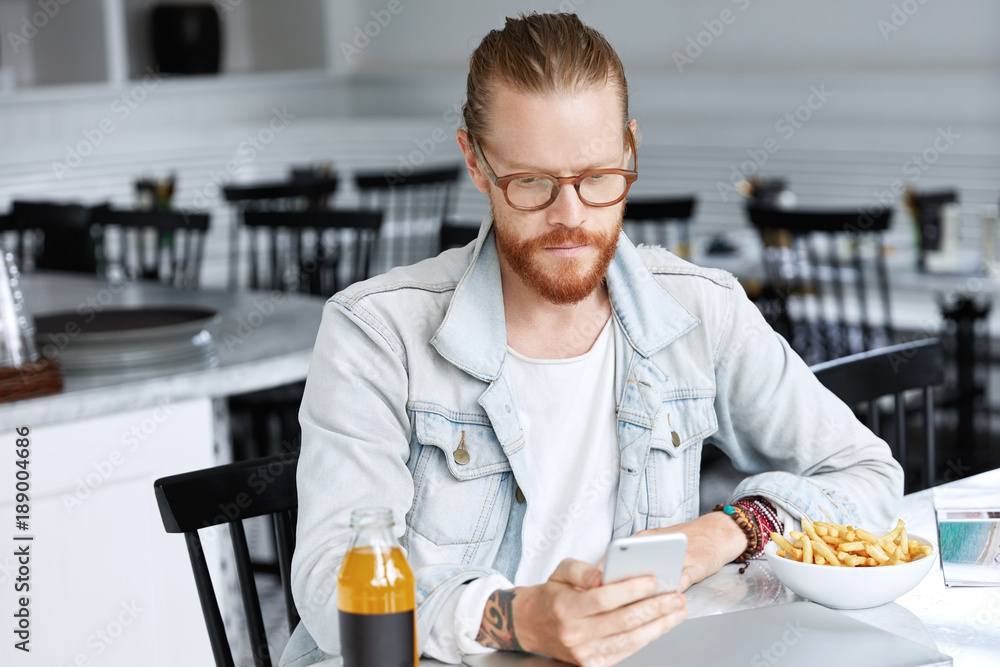 Hipster bearded male with ginger mustache, dressed in fashionable denim shirt, sits over cafe interiror, checks social networks or surfes internet websites on mobile phone during dinner break Hipster bearded male with ginger mustache, dressed in fashionable denim shirt, sits over cafe interiror, checks social networks or surfes internet websites on mobile phone during dinner break
