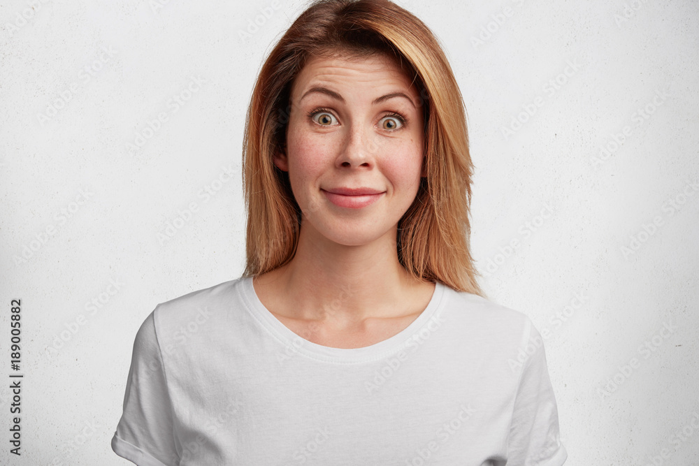 Pretty young woman with surprised pleased expression looks in bewilderment, can`t undersatand interlocutor, poses against white background. Shocked adorable female model hears intriguing news Pretty young woman with surprised pleased expression looks in bewilderment, can`t undersatand interlocutor, poses against white background. Shocked adorable female model hears intriguing news