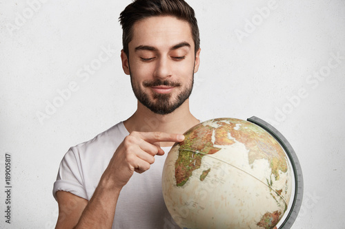 Cropped shot of young male tourist indicates at Africa continent on globe, chooses place to explore, likes travelling and go sightseeing. Handsome student studies geography indoor, locations on earth photo