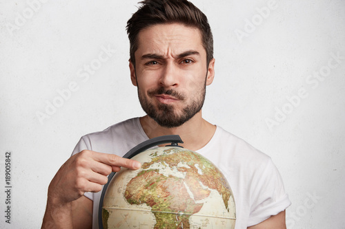 Hesitant male model with stubble indicates at globe, can`t decide where to travel, likes travelling and reaching new destinations, isolated over white background. People, tourism and lifestyle concept photo