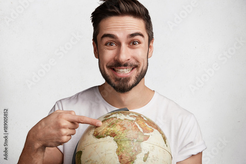 Indoor shot of smiling glad male student rejoices coming holidays, indicates at one place on globe, happy to have vacations abroad, ready exploring new places, poses against white background photo