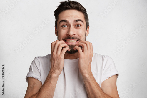 Excited pleasant looking bearded young male looks with unexpected expression at camera, clenches teeth, being nervous as sees something unbelievable, poses in studio against white background