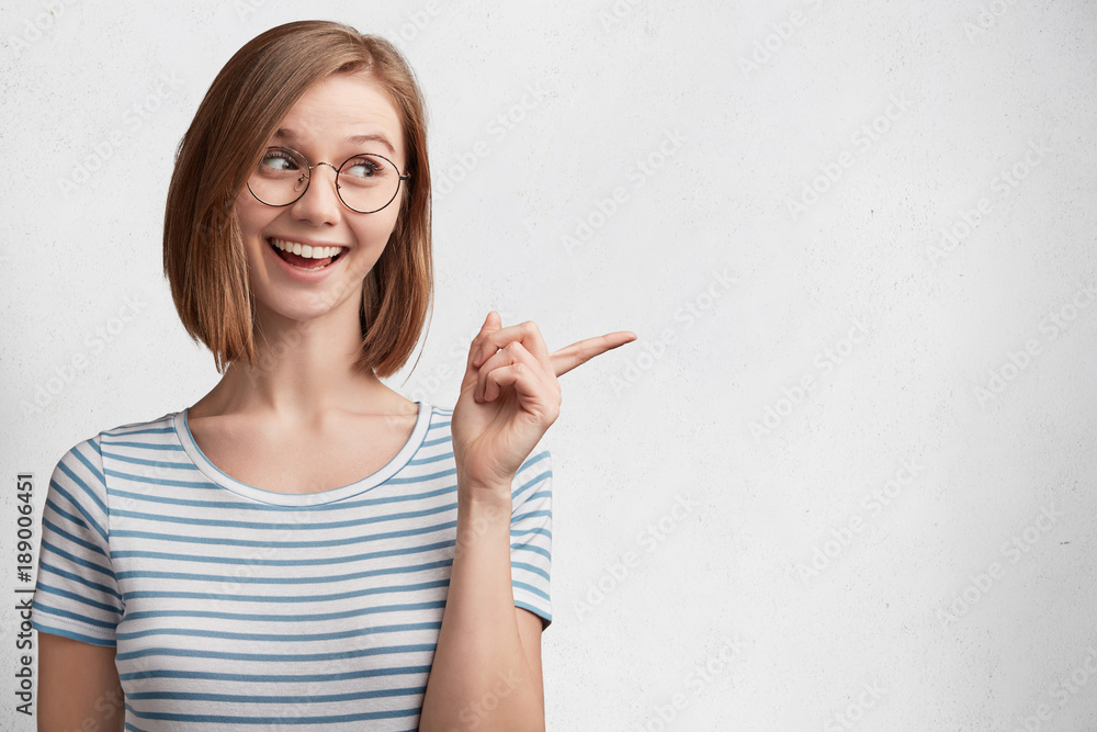 Pretty young woman dressed in striped casual t shirt, indicates with fore finger aside, demonstartes new product, poses against white background. Attractive female gestures indoor, points away Pretty young woman dressed in striped casual t shirt, indicates with fore finger aside, demonstartes new product, poses against white background. Attractive female gestures indoor, points away