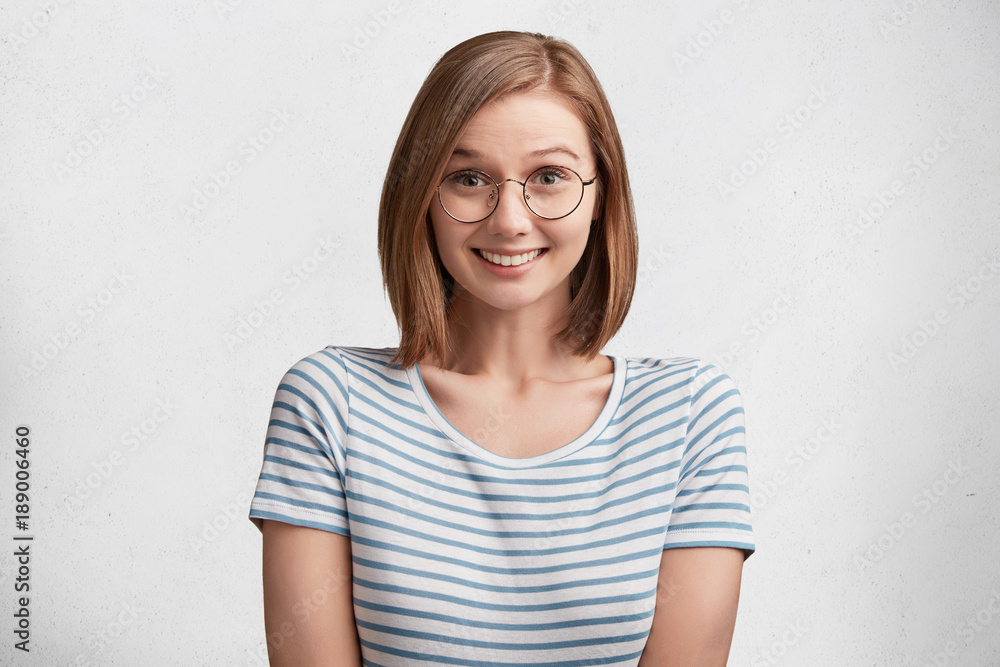 Isolated shot of beautiful young female student with happy shy expression, stands against white background, dressed casually, has appeling appearance. Woman in eyewear amazed hear pleasant news Isolated shot of beautiful young female student with happy shy expression, stands against white background, dressed casually, has appeling appearance. Woman in eyewear amazed hear pleasant news