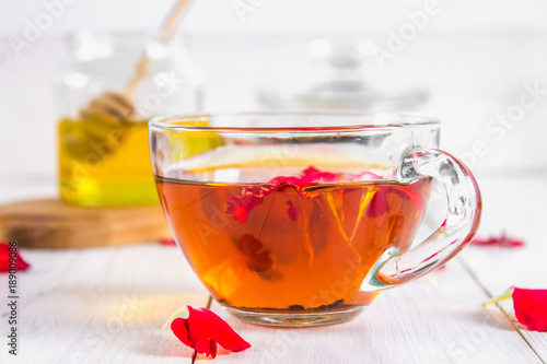 A cup of tea, in the background of a bank of honey and a jar with a black herbal floral tea on a white wooden table.