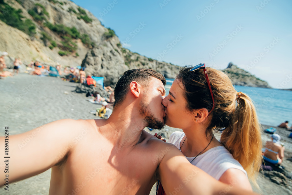 couple of travelers taking selfies and kissing on the beach on sea background couple of travelers taking selfies and kissing on the beach on sea background