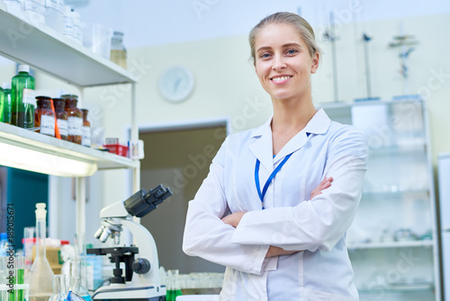 Portrait of young female scientist smiling cheerfully and looking at camera while posing in modern laboratory with arms crossed, copy space