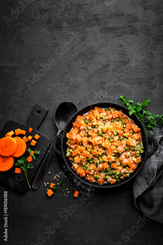 Carrot braised with fresh green peas in creamy milk sauce in stewpan, vegetable saute on black background, top view
