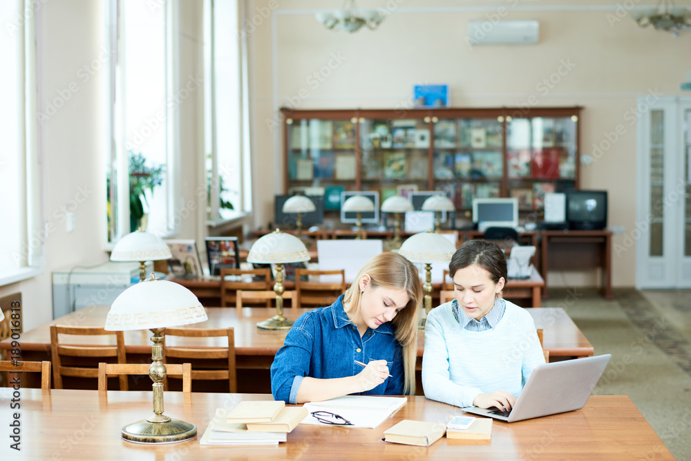 Creative college students thinking over research project while sitting at wooden table in spacious library, pretty dark-haired girl using laptop Creative college students thinking over research project while sitting at wooden table in spacious library, pretty dark-haired girl using laptop