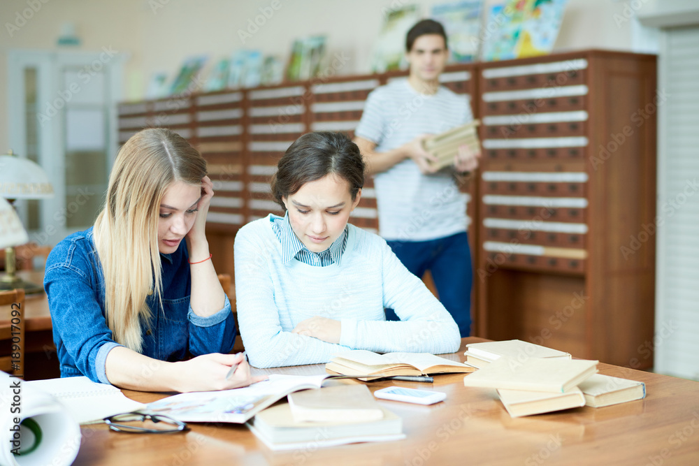 Attractive young friends sitting at desk of spacious university library and discussing their joint research project, male student with pile of books passing by Attractive young friends sitting at desk of spacious university library and discussing their joint research project, male student with pile of books passing by