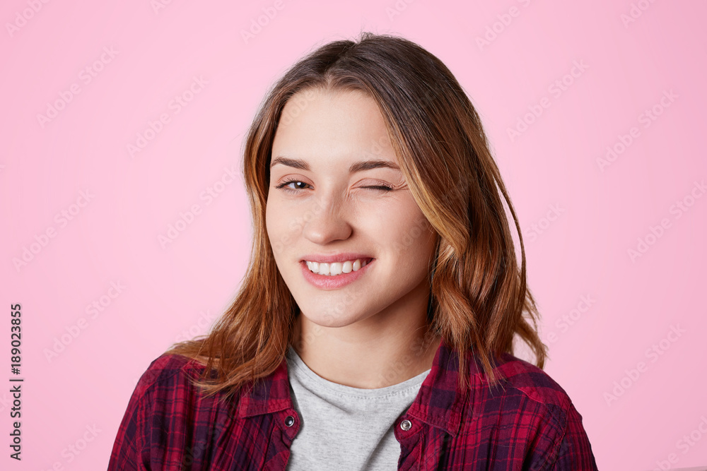 Portrait of female with cheerful expression, blinks eye as flirts with handsome man, expresses her sympathy, dressed in casual checkred shirt, isolated over pink background. Woman winks indoor Portrait of female with cheerful expression, blinks eye as flirts with handsome man, expresses her sympathy, dressed in casual checkred shirt, isolated over pink background. Woman winks indoor