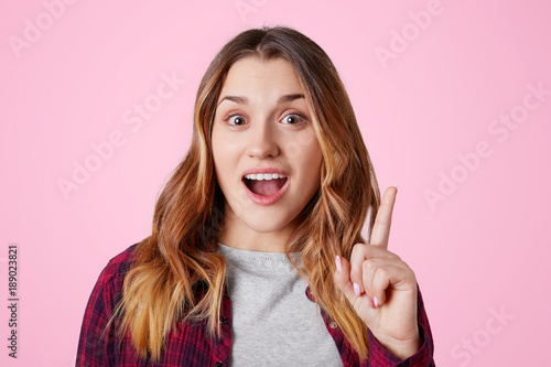 Amazed female model raises fore finger upward, keeps mouth opened, gets good idea to put in life, dressed casually, isolated over pink background. Beautiful woman remembers something important photo