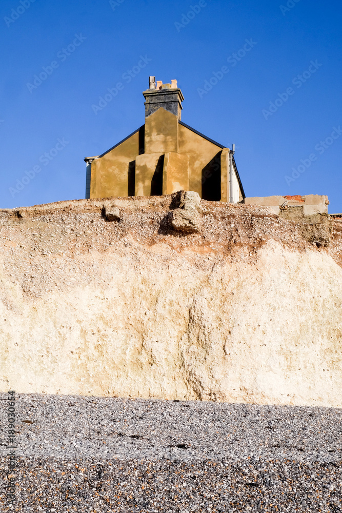 Erosion, Side of a terraced house sitting on edge of a white chalk cliff face with pebble beach beneath with eroded boulders on the beach from the cliff