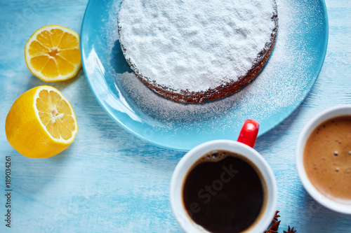 whole biscuit sprinkled with powdered sugar on the blue plate and two red cups of coffee, cut lemon and badon on blue wooden background photo