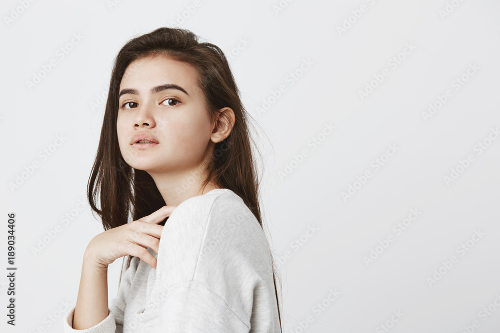 Profile of beautiful girl with dark loose hair keeping hand on breast, dressed casually, looking at camera with dark appealing eyes, posing against gray background. Beauty, youth and tenderness Profile of beautiful girl with dark loose hair keeping hand on breast, dressed casually, looking at camera with dark appealing eyes, posing against gray background. Beauty, youth and tenderness
