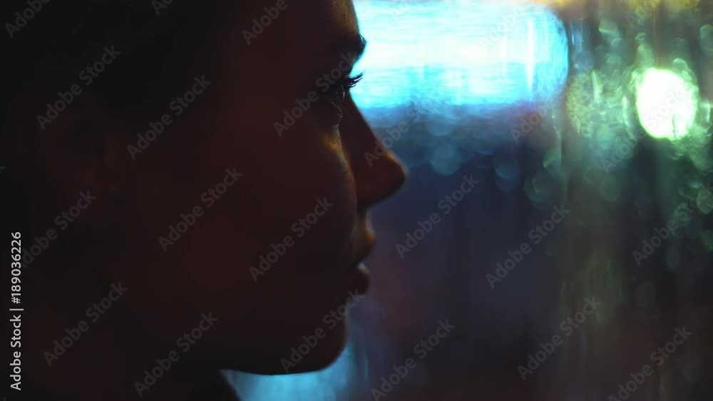 young attractive woman is sitting by the window. it is raining outside the window. large drops glide across the glass. outside the window the night and the lights of the city and passing cars. a close young attractive woman is sitting by the window. it is raining outside the window. large drops glide across the glass. outside the window the night and the lights of the city and passing cars. a close
