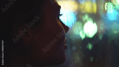 young attractive woman is sitting by the window. it is raining outside the window. large drops glide across the glass. outside the window the night and the lights of the city and passing cars. a close