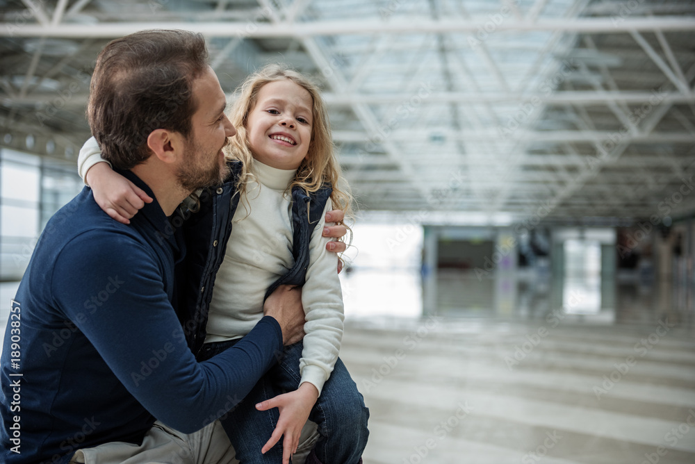 Pleased man and child standing at the airport. They are in embrace while girl is sitting on his lap. Copy space in right side