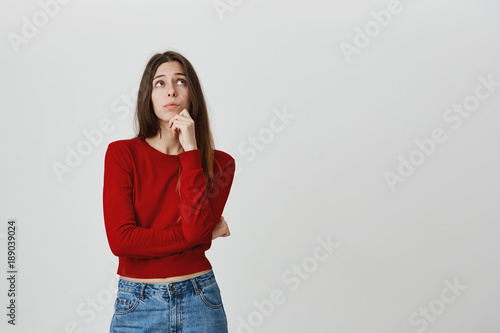 Photography Pensive dreamy student girl posing against gray background, pouting lips, looks thoughtfully upwards, touches her chin with hand