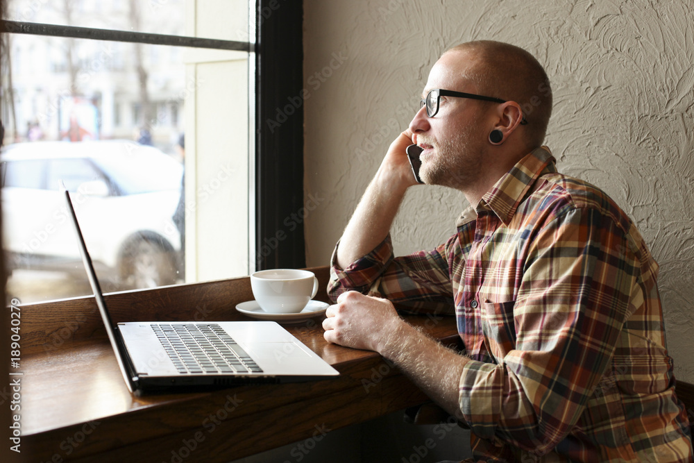 Fototapeta premium Smiling bearded businessman wearing casual hipster clothing using laptop and cell smartphone in coffe house.