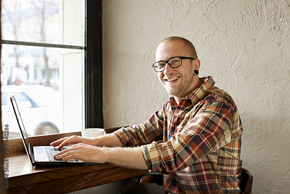 Fototapeta premium Smiling bearded businessman wearing casual hipster clothing using laptop and cell smartphone in coffe house.