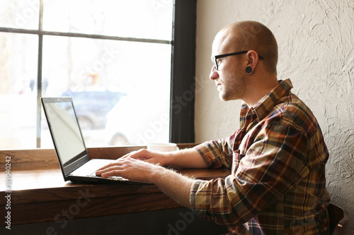 Smiling bearded businessman wearing casual hipster clothing using laptop and cell smartphone in coffe house. photo