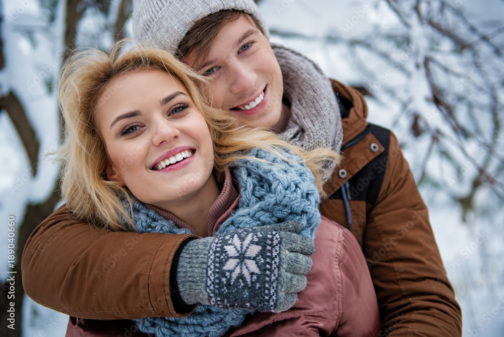 Fototapeta premium Waist up portrait of carefree young man and woman embracing and smiling. They are standing in winter forest and looking at camera with happiness
