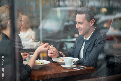 Side view smiling man telling with cheerful woman. They tasting appetizing dish while situating at desk in cafe. Dinner concept