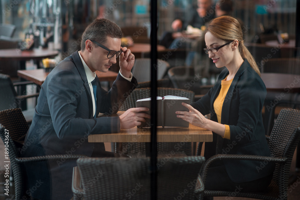 Side view cheerful male and happy girl reading information in day planner while sitting at table in cafe. Career and relax concept