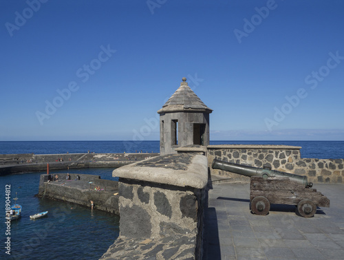 old copper cannon on paved sea front with embrasure tower, stone wall, sea horizon, fisherman ships and bathing people with clear blue sky background in Peurto de la Cruz, Tenerife