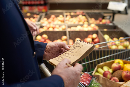 Close up of senior man holding shopping list while grocery shopping in supermarket, fruits and vegetables in background, copy space